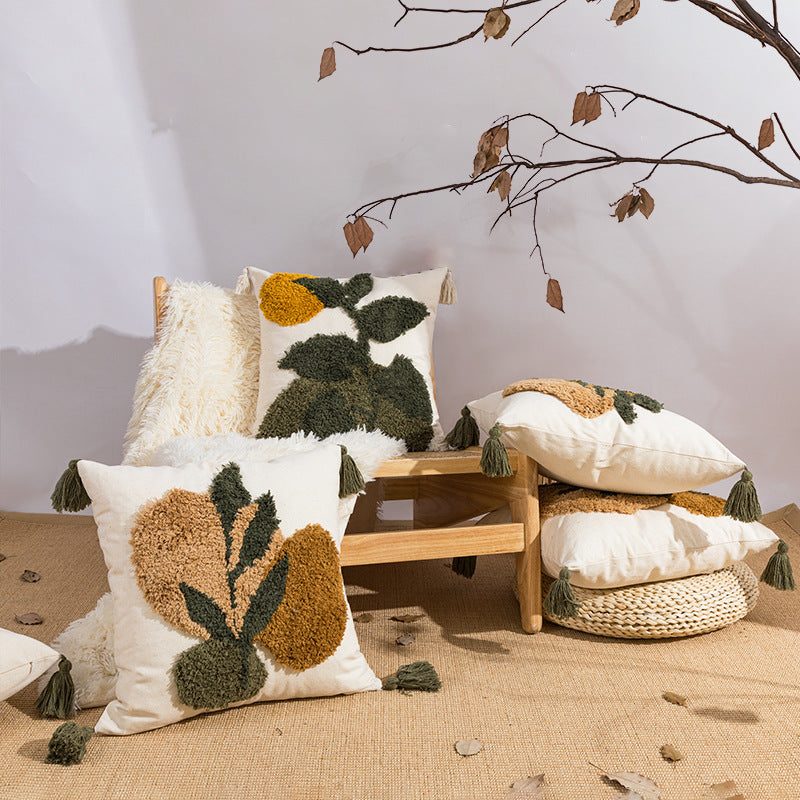 A styled arrangement of decorative throw pillows with textured botanical tufted designs in cream, olive green, and mustard yellow, displayed on a small wooden bench and floor against a neutral wall with dried branches overhead.